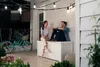 A couple sits on a white storage box on a porch at dusk, with string lights overhead and a bicycle nearby. Produktbild-Vorschau