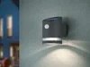 A gray outdoor wall light with a solar panel and motion sensor is mounted on a textured gray wall. The light is illuminated, casting a glow downwards. In the blurred background, a blue house with lit windows is visible. Anteprima dell’immagine prodotto