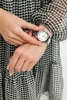 A woman's hands adjusting a silver watch on her wrist. She is wearing a black and white patterned dress. Anteprima dell’immagine prodotto