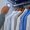 A hand is shown activating a small LED light in a closet, illuminating hanging shirts. Produktbild-Vorschau