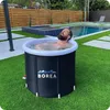 A man is relaxing in a portable ice bath in a backyard with a swimming pool and wooden fence in the background. Produktbild-Vorschau