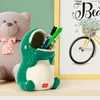A green dinosaur-shaped pen holder is shown on a shelf, with a teddy bear and a framed picture in the background. Produktbild-Vorschau