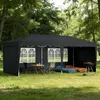 A black pop-up canopy tent set up in a grassy outdoor area with trees in the background. Inside the tent, there is a table with chairs, and sleeping mats with bedding. A grill is placed outside the tent. Náhled obrázku produktu