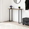A narrow console table with a wooden top and black metal legs stands against a white wall. A small vase with purple flowers and a black pot with a yellow object are on the table. A dark gray ottoman is to the right, and a rug is on the floor. Produktbild-Vorschau