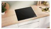 A black induction cooktop is shown on a light wood countertop, with a small potted plant to the left and kitchen accessories to the right. Produktbild-Vorschau