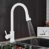 A white kitchen faucet with a pull-out spray head is shown in use, with water running into a dark sink. A framed picture and some decorative plants are visible in the background. Náhľad obrázku produktu