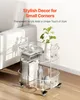A clear acrylic rolling cart with two shelves, styled with books, vases, and headphones, placed next to a white sofa. Produktbild-Vorschau