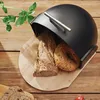 A black bread box with bread inside, placed on a wooden cutting board on a wooden table. There are also some green leaves and sliced bread in the foreground. Produktbild-Vorschau
