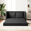 A dark gray sofa bed is shown unfolded on a light-colored patterned rug, with a large potted plant to the left and a white bookshelf to the right. Sunlight streams in from a window, creating shadows. Produktbild-Vorschau