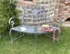 A metal garden bench with a book and a straw hat on it, set in a grassy garden with a stone wall in the background. Podgląd zdjęcia produktu