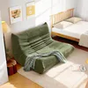 A green sofa bed is placed in a bedroom next to a wooden bed frame. Two framed pictures hang on the wall above the sofa, and a floor lamp is to the left. A blanket is draped over the sofa, and a rug is on the floor. Náhled obrázku produktu