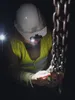 A person wearing a hard hat and a high-visibility vest is inspecting a heavy-duty chain in a dark environment, illuminated by a headlamp. Produktbild-Vorschau