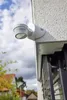 A white motion detector is mounted on the exterior wall of a building, with a cloudy sky and trees in the background. Produktbild-Vorschau