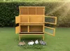 A yellow, two-story rabbit hutch with open doors, set on a green lawn with four rabbits in front. Náhľad obrázku produktu