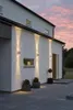 Exterior of a modern house at dusk, with wall lights illuminating the facade and pathway. Produktbild-Vorschau