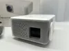 A close-up, angled view of a white and gray mini-projector on a white surface, with a blurred background showing the product's packaging. Produktbild-Vorschau