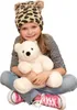 A young girl is sitting cross-legged, holding a white teddy bear. She is wearing a leopard-print hat with fuzzy ears, a striped shirt, and jeans with pink sneakers. Produktbild-Vorschau