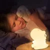 A young child sleeping in bed, holding a stuffed animal, with a dinosaur-shaped night light glowing nearby. Voir les images du produit