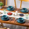 A table setting with blue ceramic dishes, cutlery, glasses of water, and small bowls of fruit, arranged on a wooden table with a white runner. Náhled obrázku produktu