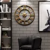 A large, vintage-style metal wall clock with Roman numerals is mounted on a gray brick wall, next to a bookshelf and a black leather armchair. Produktbild-Vorschau