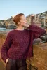 A woman wearing a purple sweater leans against a stone wall with a city and river in the background. Produktbild-Vorschau