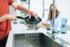 A person is rinsing a pot in a kitchen sink, with another person blurred in the background. Voir les images du produit