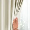 A hand is pulling back a cream-colored curtain, revealing a sheer curtain and a window with greenery outside. Podgląd zdjęcia produktu