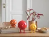 Three small, colorful, character-like figures are arranged on a wooden table, with a vase of red leaves and dried grass behind them, and a bowl of cookies to the right. Produktbild-Vorschau