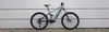 A full-suspension e-mountain bike is shown against a light gray, horizontally paneled wall. Produktbild-Vorschau