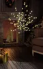 A decorative LED tree with yellow lights is placed in a cozy living room setting next to a fireplace and a sofa, with gift boxes in the foreground. Produktbild-Vorschau