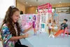 A young girl plays with a Barbie dollhouse and dolls in a bright, modern room. Produktbild-Vorschau