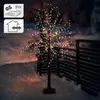 A decorative LED tree with warm white lights stands in a snowy outdoor setting at dusk, with a wooden fence in the background. Náhled obrázku produktu