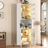 A tall white cabinet with glass doors and LED lighting, displaying various decorative items and books, situated in a well-lit room with a floor lamp and a window in the background. Produktbild-Vorschau