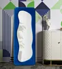 A full-length mirror with a blue velvet frame stands against a geometric patterned wallpaper. A white cabinet with a quilted texture is placed to the right of the mirror, adorned with a decorative vase and books. Podgląd zdjęcia produktu