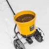 A yellow spreader with a black grate on top, sitting in the snow. The spreader has two wheels and a handle. Produktbild-Vorschau