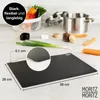 A black cutting mat is shown on a kitchen counter with ingredients like carrots and green onions in the background. Produktbild-Vorschau