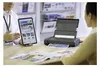 A person is showing a tablet with travel information to another person who is looking at a printer that is printing a document. Podgląd zdjęcia produktu
