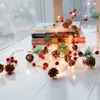 A string of Christmas lights with pinecones and red berries draped across a white surface, with books in the background. Produktbild-Vorschau