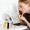 A woman applying lipstick while looking in a lighted makeup mirror, surrounded by cosmetics. Produktbild-Vorschau
