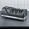A black electric knife sharpener with silver accents is placed on a dark gray surface, with a white tiled wall in the background. Podgląd zdjęcia produktu