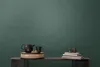 A dark green textured wall serves as the background for a table displaying a black teapot and cup on a woven tray, alongside a stack of books and decorative objects. Produktbild-Vorschau