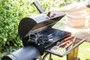 A person is opening a black BBQ grill with charcoal inside, with a basket and greenery in the background. Podgląd zdjęcia produktu