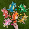 Several dragon-shaped puzzles and colorful circular game pieces are scattered on a green, grassy surface. A hand is holding one puzzle piece. Produktbild-Vorschau