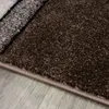 A close-up view of a brown, abstract geometric patterned rug with a beige border, lying on a light gray wooden floor. Produktbild-Vorschau
