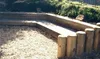 A garden setting with wooden palisades forming a raised garden bed or border, surrounded by wood chips. Produktbild-Vorschau