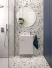 A modern bathroom with terrazzo-style tiles, a white vanity, and a round mirror. Produktbild-Vorschau