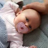 A baby lying down with a pacifier in its mouth, being petted by an adult's hand. Produktbild-Vorschau