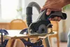 A person is using a Dremel cordless circular saw to cut a piece of wood, with a vacuum hose attached for dust collection. Produktbild-Vorschau