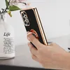 A hand holding a black phone case with a rose gold trim and a ring holder, with a vase of flowers in the background. Produktbild-Vorschau