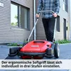 A person is using a red manual sweeper machine on a paved outdoor area next to a building. Produktbild-Vorschau